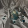 شمعة معطرة داخل كوب زجاجي بتصميم غيمة بيضاء على سطح شمع أزرق، محاطة بقلوب صغيرة، على خلفية حريرية