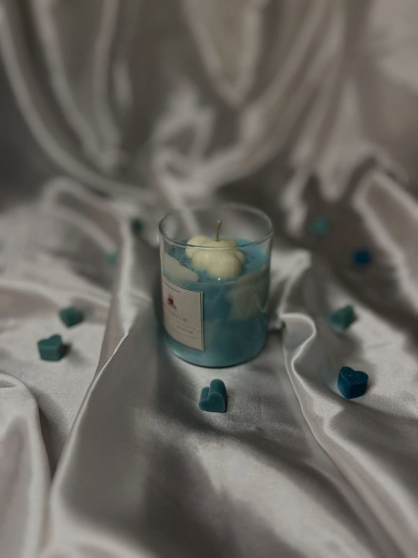 شمعة معطرة داخل كوب زجاجي بتصميم غيمة بيضاء على سطح شمع أزرق، محاطة بقلوب صغيرة، على خلفية حريرية
