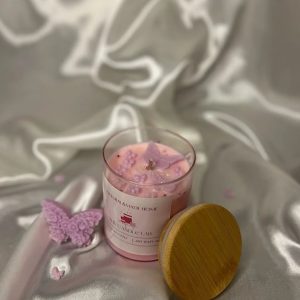شمعة وردية داخل وعاء زجاجي شفاف بغطاء خشبي، مزينة بفراشات وأشكال لؤلؤية بلون بنفسجي، موضوعة على قماش حريري عاجي