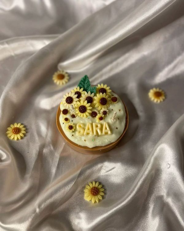 شمعة على شكل كيكة مزينة بزهور دوار الشمس، مكتوب عليها اسم "SARA"، موضوعة على قماش حريري بلون عاجي