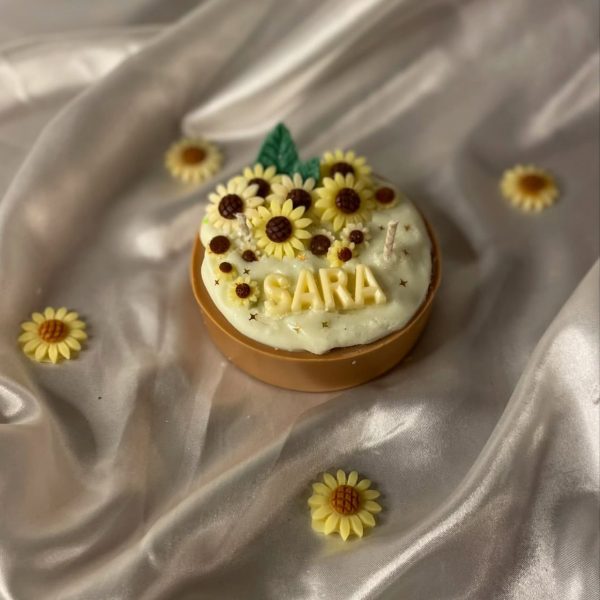 شمعة على شكل كيكة مزينة بزهور دوار الشمس، مكتوب عليها اسم "SARA"، موضوعة على قماش حريري بلون عاجي