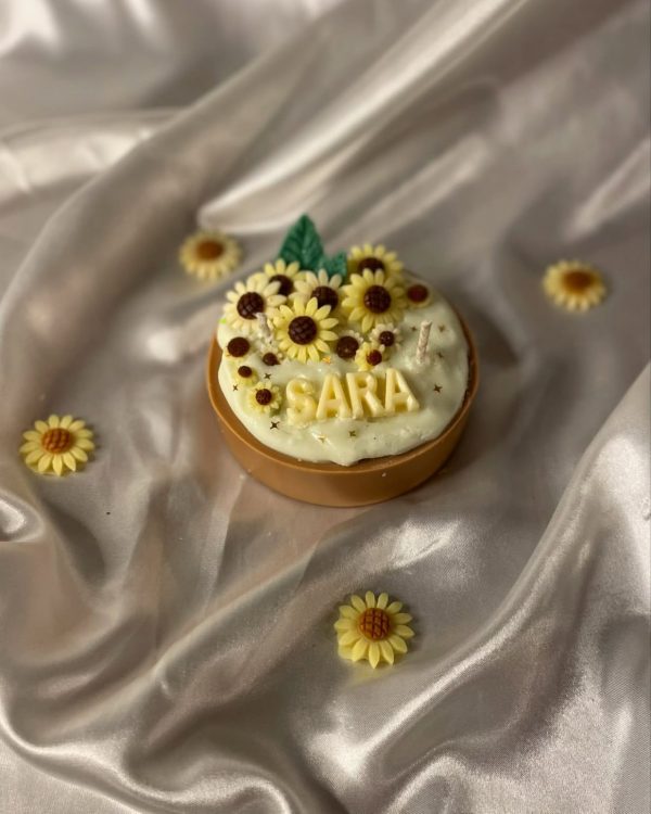 شمعة على شكل كيكة مزينة بزهور دوار الشمس، مكتوب عليها اسم "SARA"، موضوعة على قماش حريري بلون عاجي