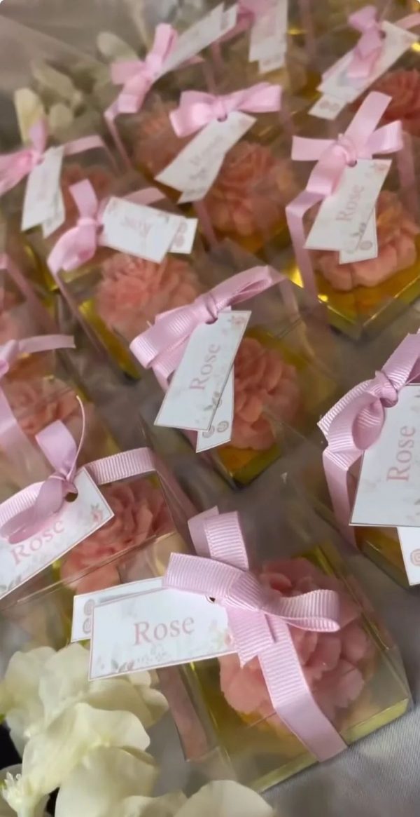 توزيعات شموع بتصميم وردة وردية داخل علبة شفافة مربوطة بشريط وردي وبطاقة تحمل كلمة "Rose".