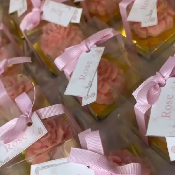 توزيعات شموع بتصميم وردة وردية داخل علبة شفافة مربوطة بشريط وردي وبطاقة تحمل كلمة "Rose".