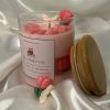 شمعة وردية داخل وعاء زجاجي شفاف، مزينة بزهرة توليب وقلوب صغيرة وفراشة بيضاء، موضوعة على قماش ساتان أبيض لامع