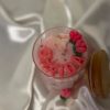 شمعة وردية داخل وعاء زجاجي شفاف، مزينة بزهرة توليب وقلوب صغيرة وفراشة بيضاء، موضوعة على قماش ساتان أبيض لامع