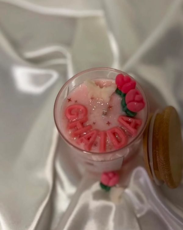 شمعة وردية داخل وعاء زجاجي شفاف، مزينة بزهرة توليب وقلوب صغيرة وفراشة بيضاء، موضوعة على قماش ساتان أبيض لامع