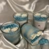 شموع زرقاء بديكورات غيوم وكلمات مثل "BABY" داخل أوعية زجاجية شفافة، موضوعة على قماش حريري أبيض