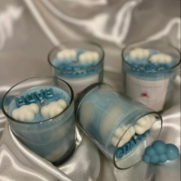 شموع زرقاء بديكورات غيوم وكلمات مثل "BABY" داخل أوعية زجاجية شفافة، موضوعة على قماش حريري أبيض