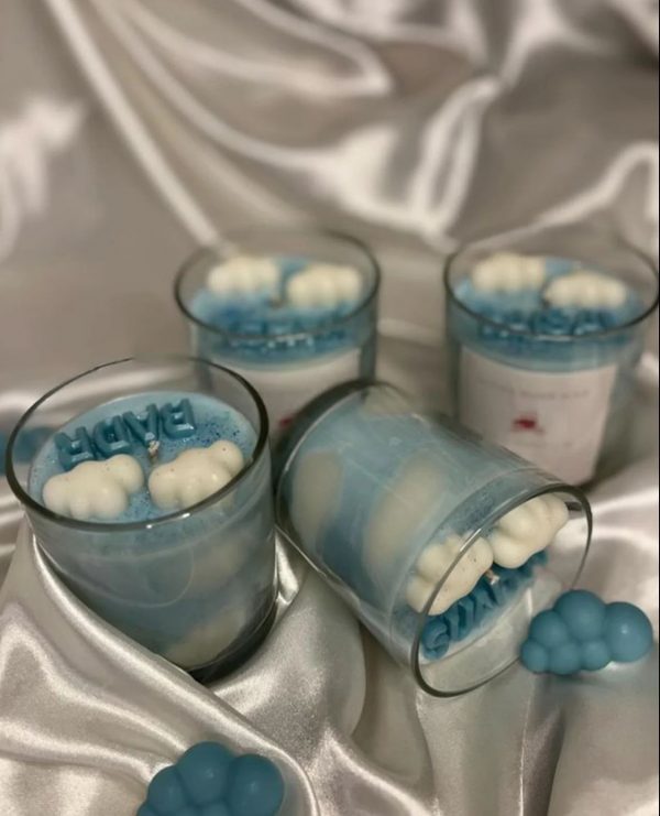 شموع زرقاء بديكورات غيوم وكلمات مثل "BABY" داخل أوعية زجاجية شفافة، موضوعة على قماش حريري أبيض