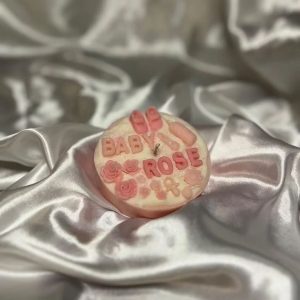شمعة صغيرة على شكل كيك بلون وردي فاتح مزينة بزهور صغيرة وكلمة "BABY ROSE"، موضوعة على قماش ساتان لؤلؤي ناعم.