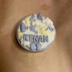 شمعة كيك بيضاء مزينة بعناصر المواليد باللون الأزرق والأصفر، وتحمل اسم "KENAN"، موضوعة على ورق تغليف بني ناعم.