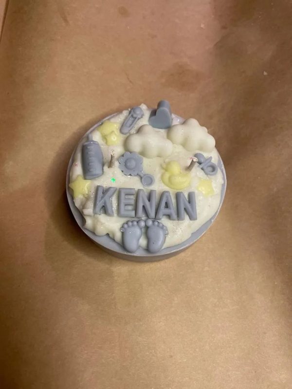 شمعة كيك بيضاء مزينة بعناصر المواليد باللون الأزرق والأصفر، وتحمل اسم "KENAN"، موضوعة على ورق تغليف بني ناعم.